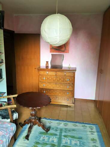 A cozy room featuring a round wooden table, an ornate chest of drawers, a large paper lantern, pale pink walls, a blue patterned rug, and wooden furniture.