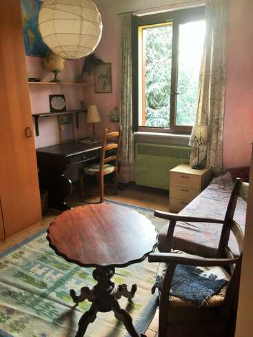 Cozy room with pastel pink walls, wooden furniture, and large window. Features a round table, armchair, desk with a globe, lamp, and decorative rug.