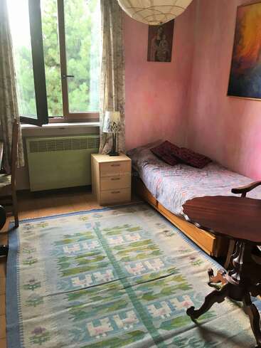 A cozy, small bedroom with a single bed, pink walls, patterned rug, wooden furniture, open window, floral curtains, lamp, and colorful wall art.