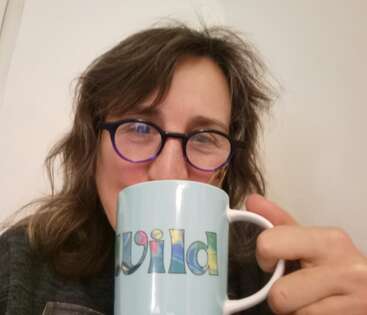 Une femme à lunettes et aux cheveux ondulés sourit en tenant un mug bleu clair sur lequel est écrit le mot "wild" en lettres colorées.