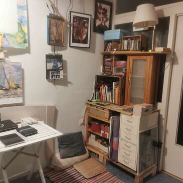 Un coin bureau confortable comprend une table blanche, des étagères, diverses œuvres d'art, des calendriers, des coussins, une lampe de bureau, des livres et des articles de papeterie organisés dans un espace compact.