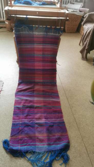 Un métier à tisser manuel avec un textile vibrant, tissé dans des tons de violet, de bleu et de rouge, s'étend jusqu'au sol. Les extrémités sont frangées et l'espace de travail est confortable et créatif.