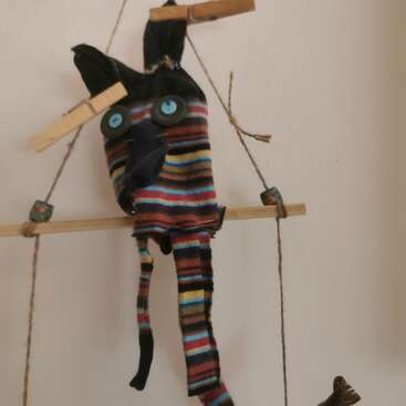 Une marionnette faite à la main, avec des yeux en forme de boutons et un tissu rayé coloré, est suspendue à une tige en bois, maintenue par des pinces à linge et de la ficelle, contre un mur ordinaire.