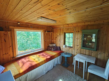 Cette chambre en bois chaleureuse comprend un lit simple recouvert d'une couverture colorée, une grande fenêtre, des petites tables, une chaise blanche et une armoire verte ornée d'objets décoratifs.