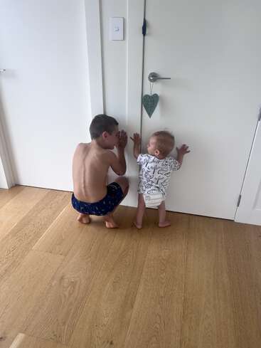 A shirtless boy and a toddler in a diaper are quietly listening at a closed white door on a wooden floor, appearing curious and playful together.