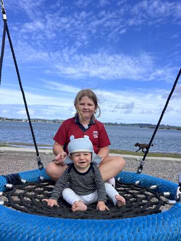 A young girl and a baby sit together on a large swing by the water, smiling under a blue sky, with a dog in the background.