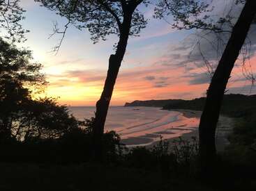 The image depicts a serene beach scene at sunset, featuring a sandy shore, gentle waves, and lush trees silhouetted against a vibrant sky with hues of orange, pink, and blue.