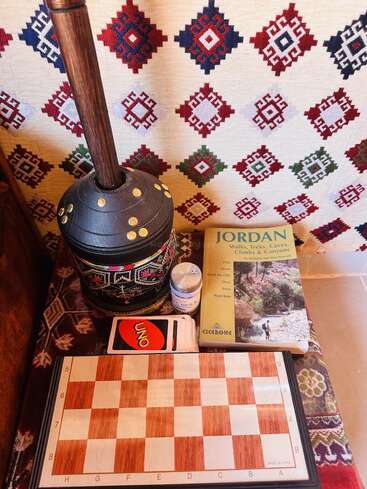 Une installation confortable comprenant une tapisserie à motifs, un tambour traditionnel, un guide "Jordanie", des cartes UNO, un petit récipient et un échiquier en bois sur un tissu décoratif.