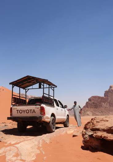 Un homme se tient à côté d'un pick-up Toyota blanc dans un paysage désertique sablonneux, avec des falaises rocheuses en arrière-plan, sous un ciel bleu clair et lumineux.