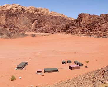 L'image montre un camp isolé dans le désert avec plusieurs petits bâtiments sur du sable rouge, entouré de falaises rocheuses escarpées sous un ciel bleu lumineux.