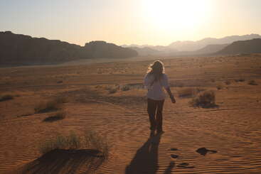 Une personne marche seule dans un vaste paysage désertique éclairé par le soleil. Les dunes de sable, les plantes éparses et les montagnes lointaines créent une atmosphère sereine, solitaire et paisible.