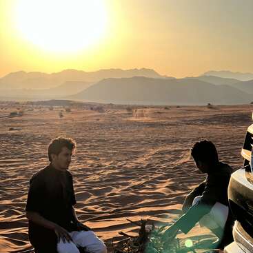 Dos hombres sentados en la cálida arena del desierto junto a un vehículo, con las montañas y el sol poniente de fondo, creando una escena pacífica y contemplativa al atardecer.