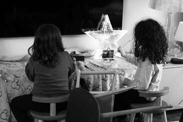 Two children sit at a table, their backs turned, engaged in arts and crafts. A glowing star-shaped light decorates the cozy, softly lit room.