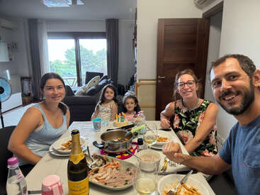 A family of five, including two children, enjoys a meal together indoors. There’s food, drinks, a birthday cake with candles, and everyone is smiling happily.