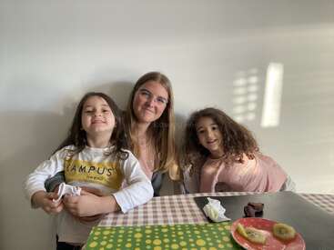 Three people, two children and one adult, pose happily at a table. On the table are slices of kiwi, an orange, brownies, and a napkin.