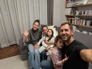 A happy family of five sits on a couch, smiling for a selfie. One woman waves, while shelves with books and photos fill the cozy background.