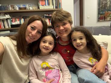 Four people sit closely together on a couch, smiling at the camera. Two adults and two young girls, both girls wearing matching Princess Peach shirts. Cozy atmosphere.