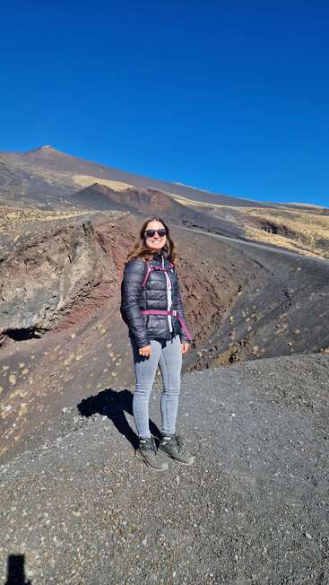 A woman stands smiling on rocky volcanic terrain, wearing sunglasses, a black jacket, grey pants, and hiking boots under a clear blue sky, mountainous landscape behind.