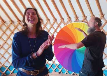 Dos personas interactúan alegremente dentro de una yurta, de pie frente a una obra de arte circular de vivos colores. Ambas parecen estar riendo y disfrutando del momento.