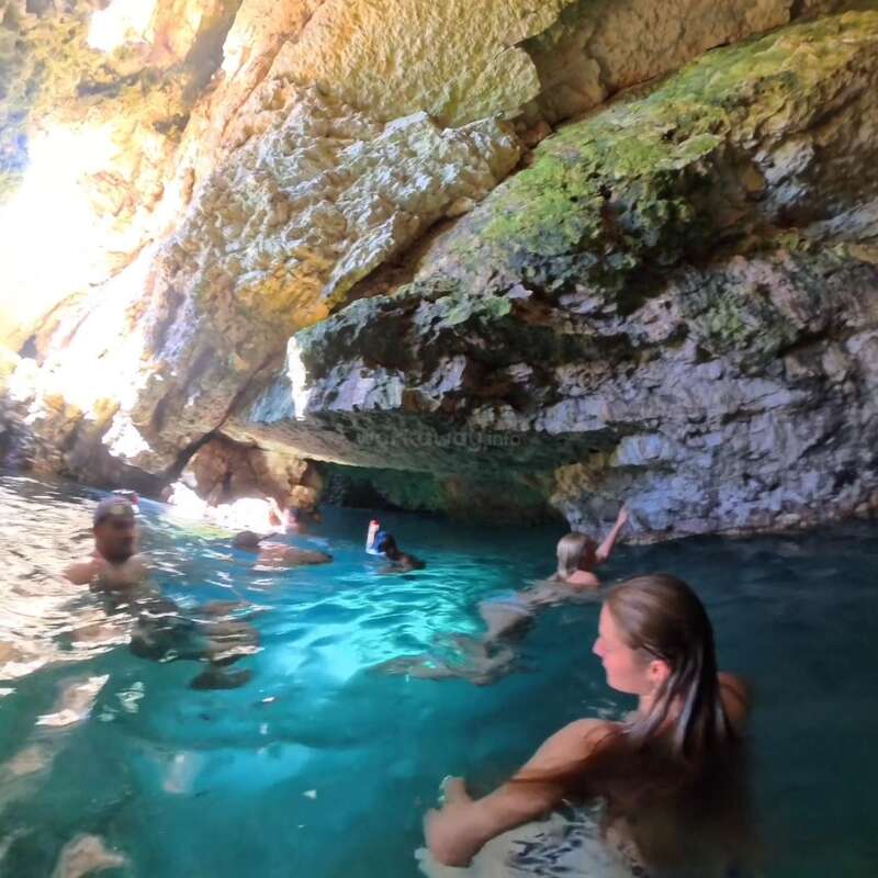 Várias pessoas nadam e mergulham com snorkel nas águas azuis e cristalinas dentro de uma caverna rochosa, iluminada pela luz natural do sol que entra pelas aberturas no teto e nas paredes da caverna.