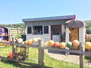 A quaint seaside ice cream shack decorated with colorful buoys and wooden fencing, offering snacks while surrounded by green hills under a clear, sunny blue sky.