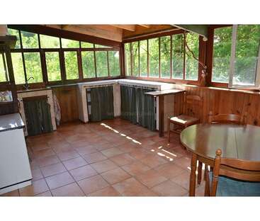 This sunroom kitchen features large windows, wooden walls, tiled floor, a round dining table, simple cabinetry with curtains, and abundant natural light overlooking green scenery.