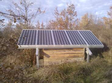 The image shows a large solar panel installed on top of a small wooden shed, surrounded by tall grass and trees with autumn-colored leaves.