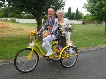 Das Bild zeigt ein älteres Ehepaar, das auf einem gelben, mit Blumen geschmückten Dreirad auf einer Straße in einem parkähnlichen Umfeld fährt und damit ein Gefühl von Freude und Festlichkeit vermittelt.