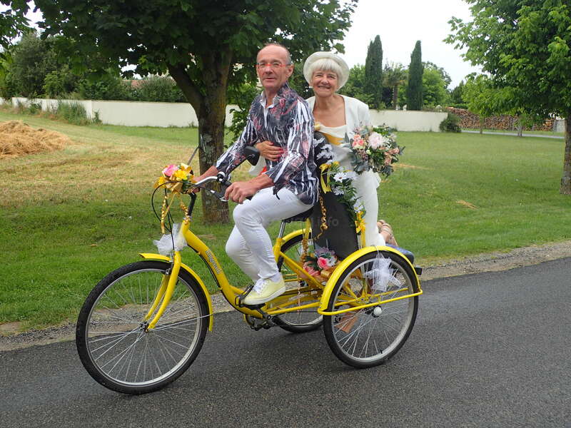The image depicts an elderly couple riding a yellow tricycle, adorned with flowers, on a road in a park-like setting, conveying a sense of joy and celebration.