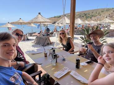 Une famille de cinq personnes est assise dans un restaurant au bord de la mer, en train de boire un verre. La journée ensoleillée, la mer bleue et les parasols de paille créent une atmosphère de vacances détendue.