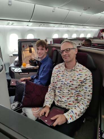 Deux personnes sont assises dans une luxueuse cabine d'avion et sourient à la caméra. L'une porte une chemise colorée avec des avions, tandis que l'autre prend un jus d'orange et un oreiller.