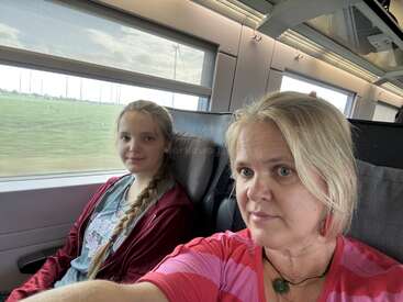 Une femme et une fille sont assises ensemble dans un train et regardent la caméra. Des champs verdoyants et des éoliennes sont visibles par la fenêtre derrière elles.