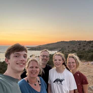 Five people pose happily for a group selfie outdoors at sunset, with a scenic coastal landscape, calm sea, and hills in the background. Warm, peaceful atmosphere.