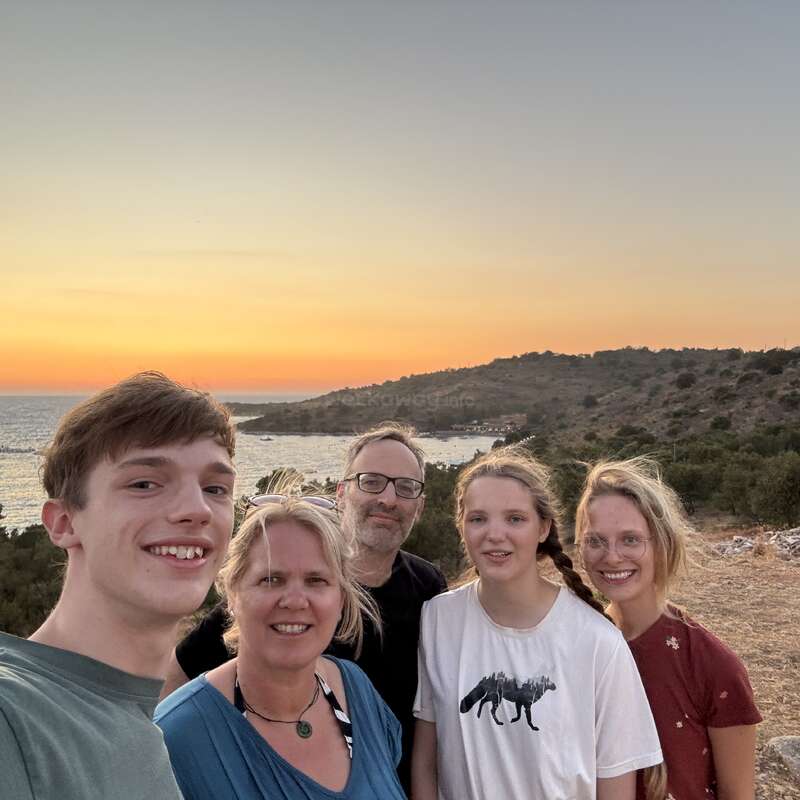 Cinq personnes posent joyeusement pour un selfie de groupe en plein air au coucher du soleil, avec un paysage côtier pittoresque, une mer calme et des collines en arrière-plan. Ambiance chaleureuse et paisible.