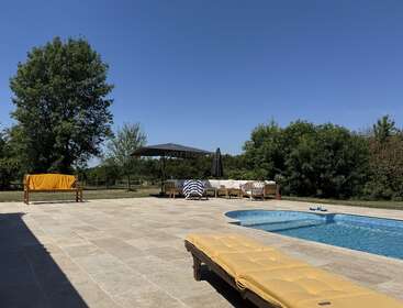 Uma área de piscina ao ar livre ensolarada com azulejos bege, espreguiçadeiras, uma toalha amarela, guarda-sóis e árvores verdes exuberantes ao fundo, sob um céu azul claro.