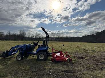 Um trator azul com um cortador de grama vermelho está parado em um campo gramado sob um céu parcialmente nublado, com o sol brilhando forte acima. Árvores se alinham ao fundo.