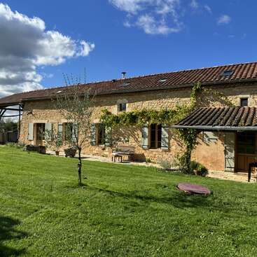 Ein hübsches Bauernhaus aus Stein mit Ziegeldach, umgeben von grünem Gras, Kletterpflanzen, Bäumen, Bänken und blauem Himmel mit Wolken an einem sonnigen Tag.