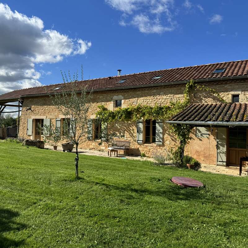 Uma charmosa casa de fazenda de pedra com telhado de telhas, cercada por grama verde, trepadeiras, árvores, bancos e céu azul com nuvens em um dia ensolarado.