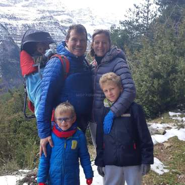 La imagen muestra a una familia de cinco miembros posando para una foto ante un fondo montañoso, con el padre llevando un bebé a la espalda.
