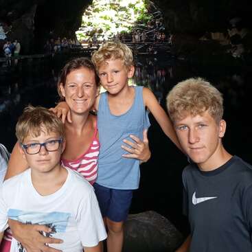 Une femme souriante et trois garçons posent ensemble à l'intérieur d'une grotte remplie d'eau, la lumière du soleil filtrant par l'entrée, tandis que des plantes vertes sont visibles à l'arrière-plan.