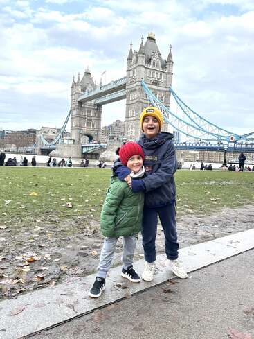 Zwei fröhliche Kinder in bunten Winterjacken und Mützen stehen an einem bewölkten Tag vor der berühmten Tower Bridge in London und haben Spaß.
