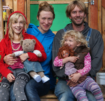 Una feliz familia de cuatro miembros está sentada junta, sonriendo cálidamente. Los niños sostienen muñecas y llevan trajes coloridos, mientras que los padres los abrazan con cariño en un ambiente acogedor.