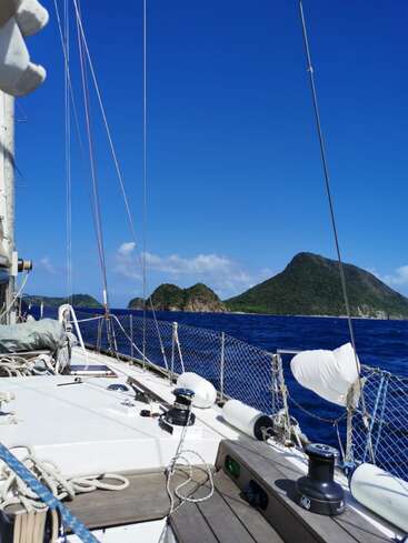 The image depicts a serene scene of a white sailboat on the open ocean, with a majestic mountainous island visible in the distance under a clear blue sky.