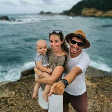The image depicts a family of three posing on a rocky beach, with the man wearing a hat and sunglasses, the woman holding a baby, and the ocean serving as a serene backdrop.