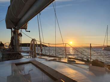 The image depicts a serene sunset scene from the deck of a sailboat, with the sun setting over the water and a clear blue sky.