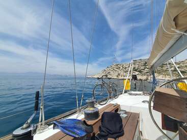 The image depicts a serene scene of a sailboat on the water, with a bicycle and other items on board, set against a picturesque rocky coastline and blue sky.