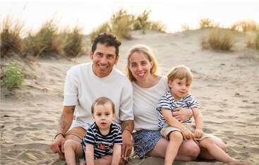A happy family of four poses on a sandy beach. Both parents smile warmly while holding their two young sons. Tall grass grows in the background.