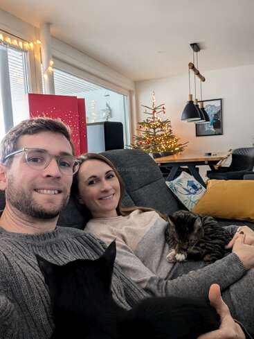 A couple sits on a cozy couch with two cats, smiling. Behind them is a decorated Christmas tree, festive lights, and a modern, inviting living room.
