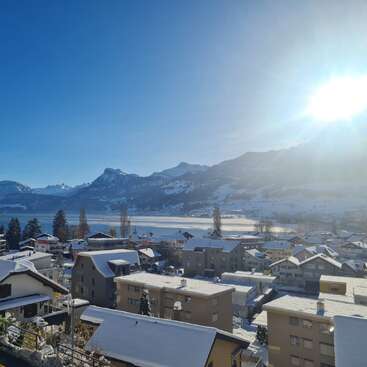 A serene winter scene of a snowy village beneath clear blue skies. Majestic mountains and a bright sun overlook the peaceful houses and shimmering frozen lake.