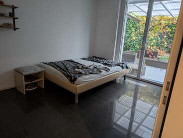 Une chambre à coucher minimale avec un matelas sur un cadre de lit beige, une literie grise, une petite table de nuit en bois, des étagères murales et de grandes portes vitrées donnant sur un patio vert et verdoyant.